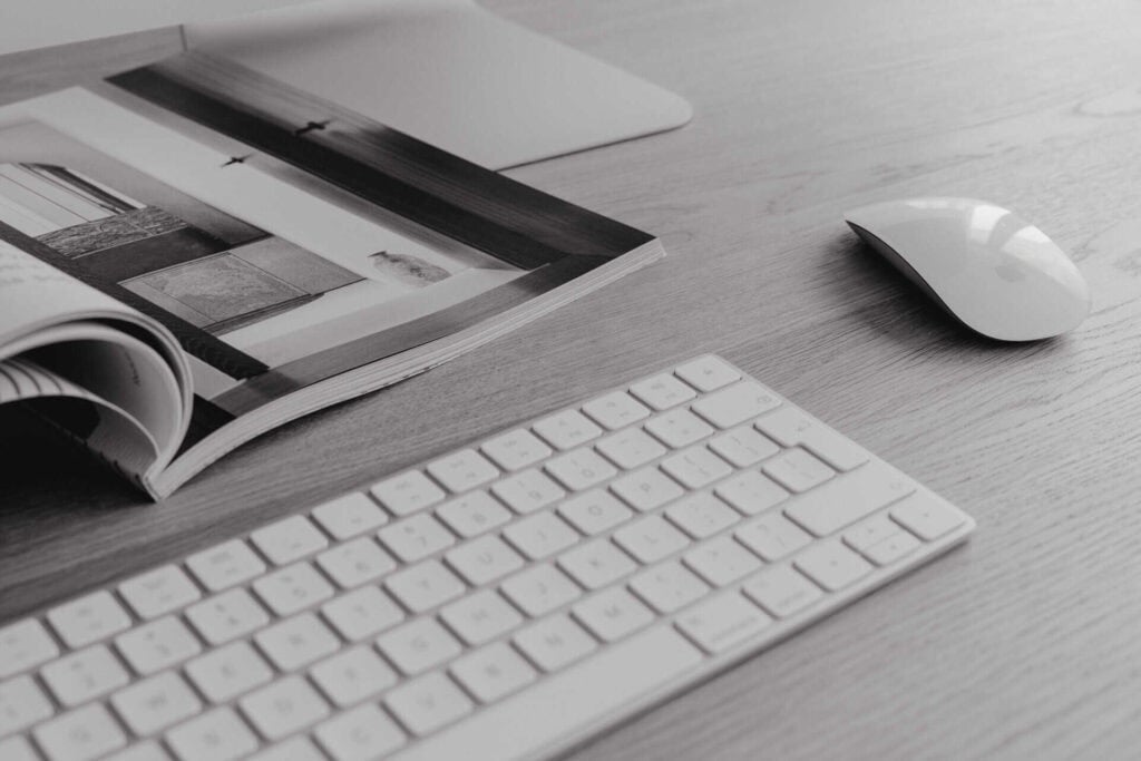 A clean and minimalistic workspace featuring a white keyboard, an Apple Magic Mouse, and an open magazine displaying architectural images. The setup is arranged on a light wooden desk, creating a sleek and modern aesthetic.
