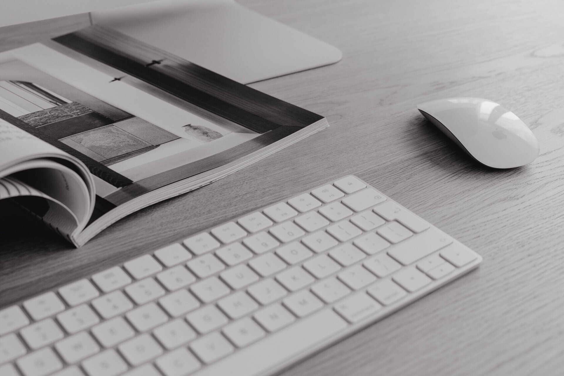 A clean and minimalistic workspace featuring a white keyboard, an Apple Magic Mouse, and an open magazine displaying architectural images. The setup is arranged on a light wooden desk, creating a sleek and modern aesthetic.